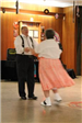 A man wearing suspenders dances with a woman in an orange skirt. 